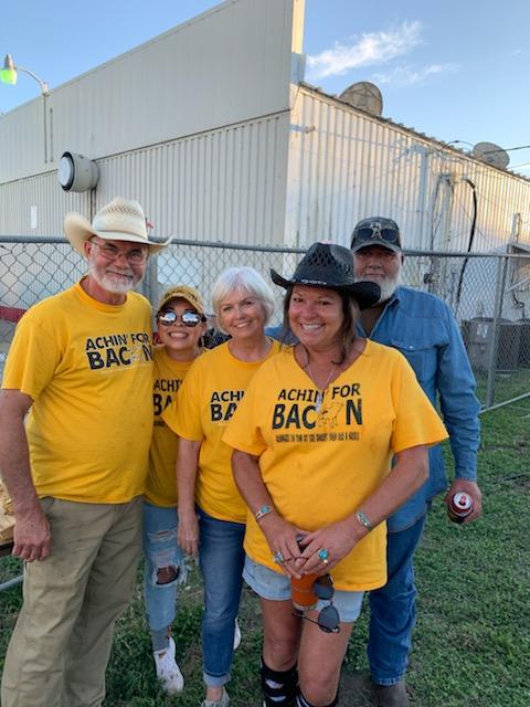 A group of 5 people stands smiling for the camera wearing yellow shirts that say "Achin' for Bacon."