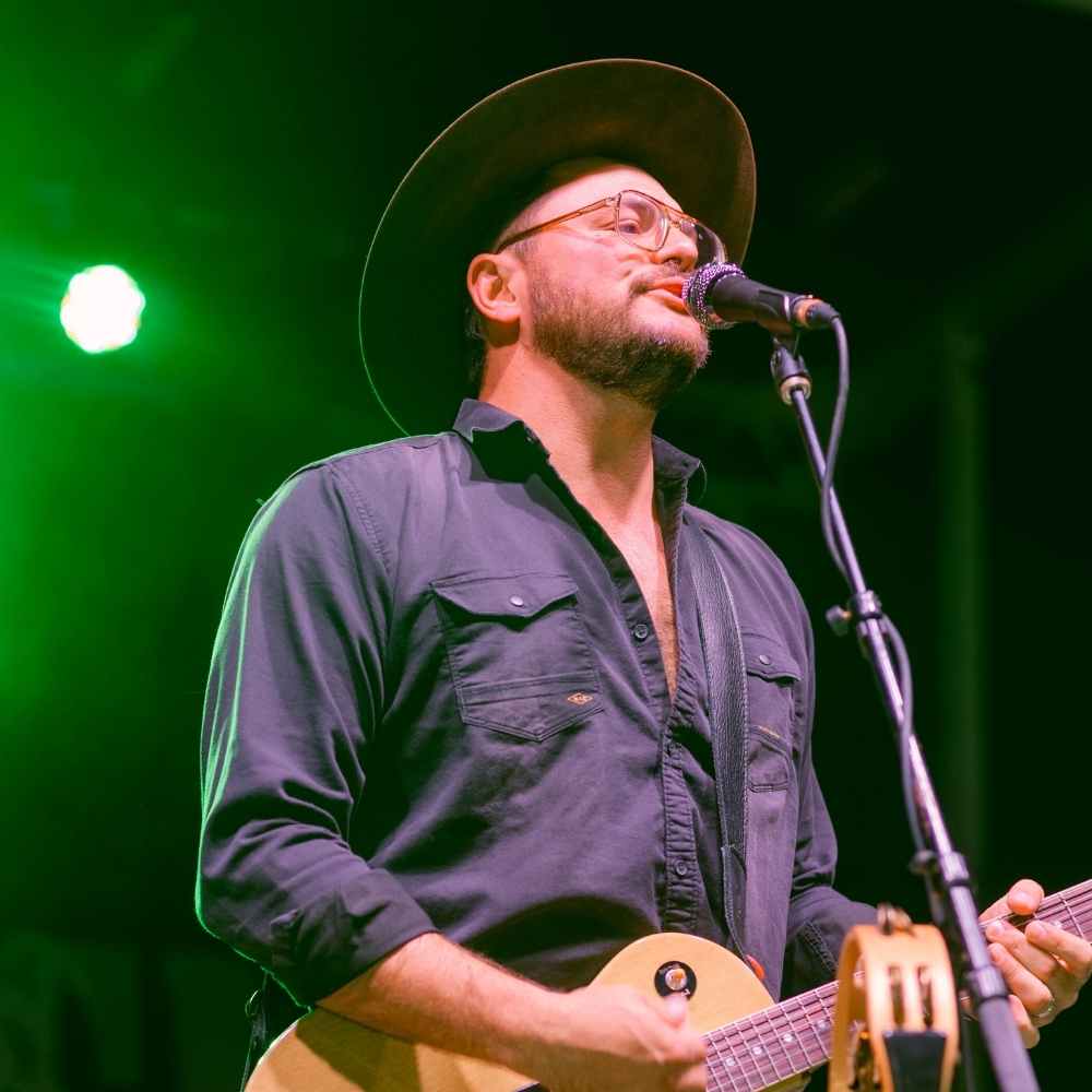 Josh Weathers sings live at Bacon Bash Texas wearing glasses, a dark shirt, and a wide-brim hat performs on stage under green lighting while singing into a microphone and playing guitar.