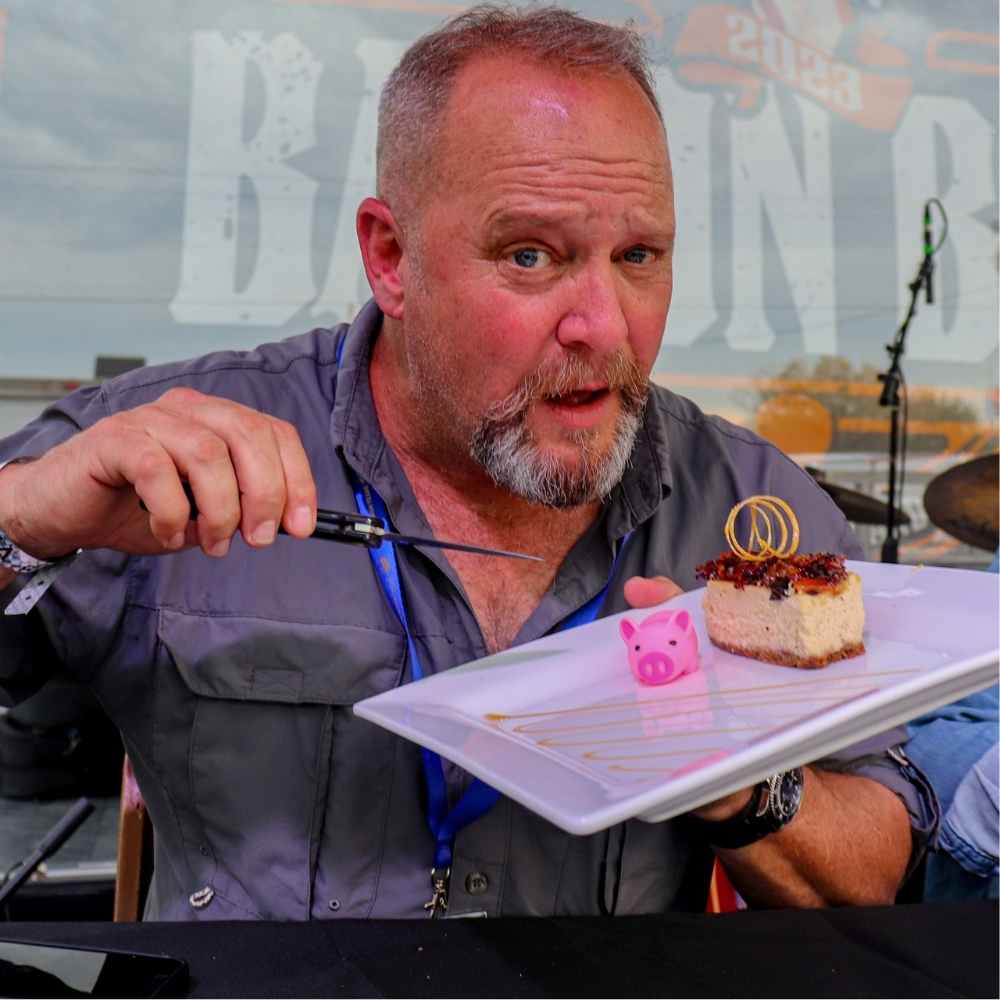 Jeff Tucker, final table judge at Bacon Bash Texas, wearing a collared shirt dramatically holds a plated bacon-themed dessert topped with garnish, with a small pink pig figurine on the plate.